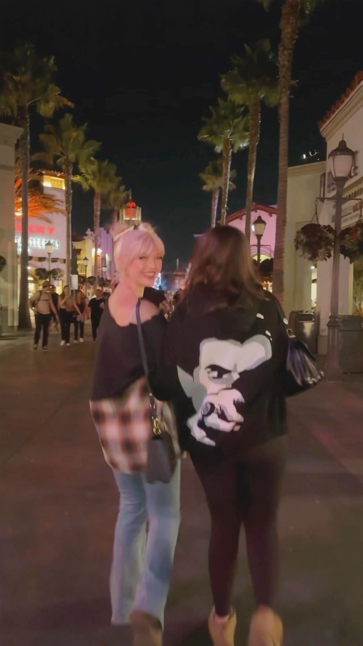 two women are walking on a street at night, flanked by palm trees and buildings. the woman on the left is wearing a black top, a red and pink flannel shirt, and blue jeans. the woman on the right is wearing a black sweatshirt with a large face on the back and black leggings. a group of people is walking behind the women in the background.