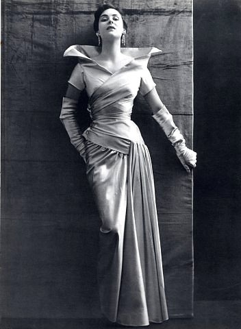 a black and white portrait of a woman in a glamorous dress. she is posing in a studio setting with a backdrop of textured fabric. the woman is wearing a flowing gown with a dramatic neckline and long, fitted sleeves. she has a classic hairstyle and makeup, with a touch of jewelry to complete the look.