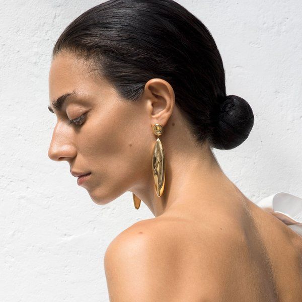 a woman is shown in profile, wearing large gold earrings. her hair is pulled back into a bun. the focus is on her face and the jewelry. her shoulder and back are visible.