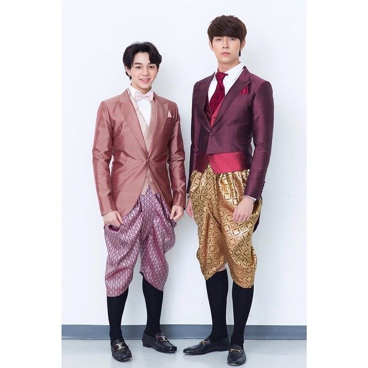 two men are standing and smiling. they are both wearing traditional thai clothing, which includes a suit jacket, patterned pants, black socks, and black dress shoes. the man on the left is wearing a pink suit jacket and pink pants, while the man on the right is wearing a burgundy suit jacket and gold pants.