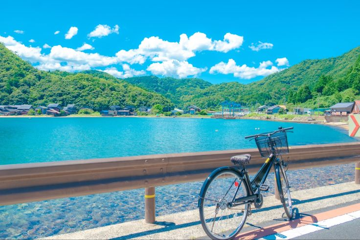 a bicycle is parked on a roadside railing overlooking a picturesque scene of a clear blue lake surrounded by green mountains and a small village under a bright blue sky with fluffy white clouds.
