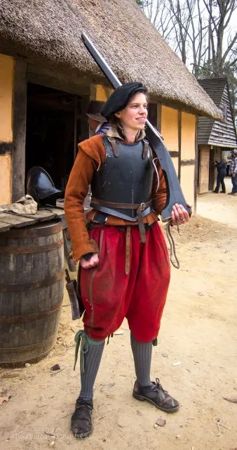 a man dressed as a medieval soldier is standing outside a wooden building. he is holding a musket in his left hand and has a serious expression on his face. the man is wearing plate armor, a dark red belt, and a black hat. he is standing on dirt ground in front of a thatched roof building. there are trees in the background.