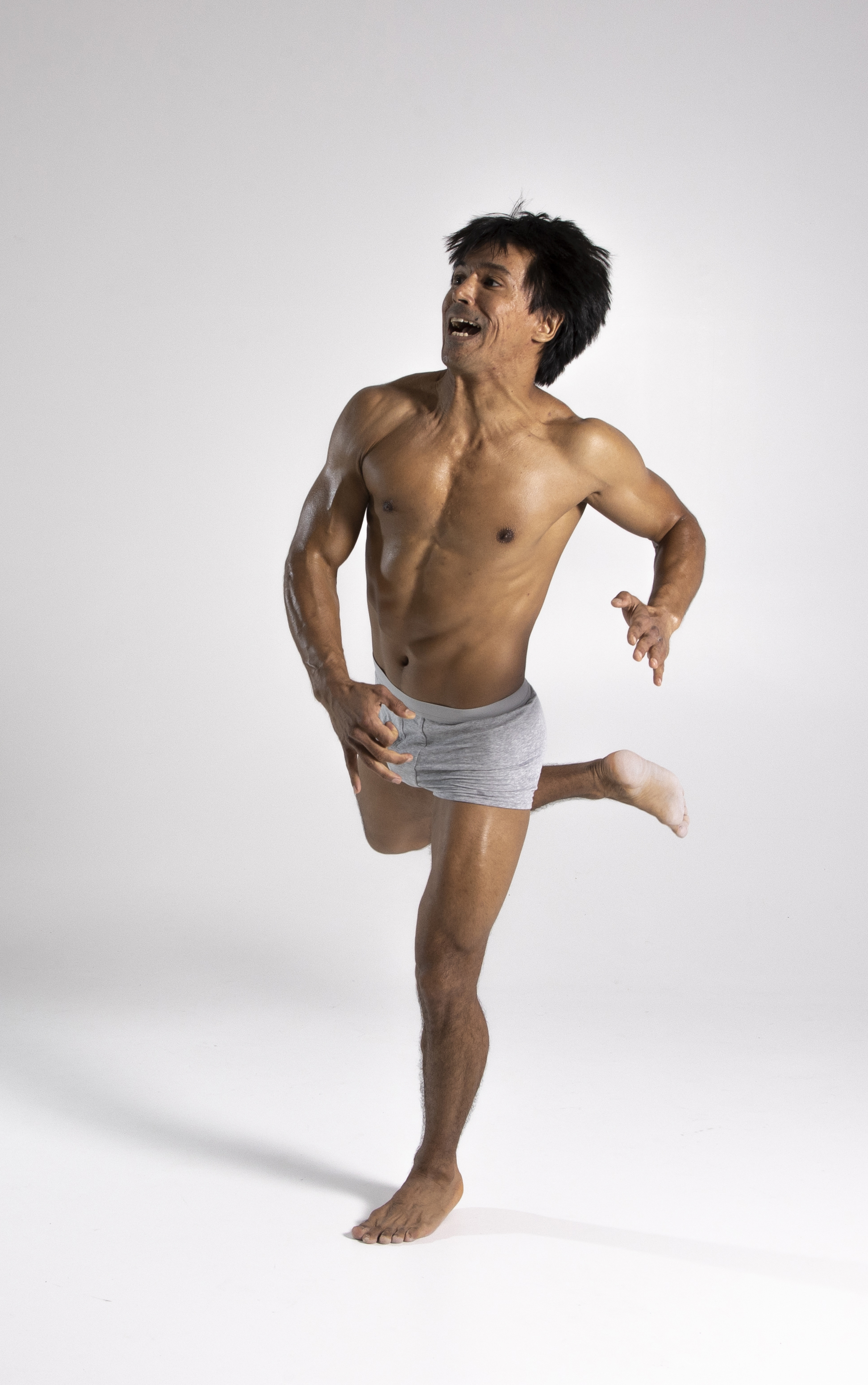 a muscular, shirtless man wearing underwear is jumping in a studio shot. he is barefoot and smiling. he appears to be in athletic shape.