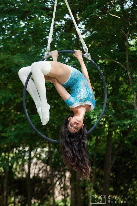 a woman in a blue and white outfit is hanging upside down from an aerial hoop in a forest setting. the hoop is suspended from the trees above her, and she is holding onto it with her arms and legs. her long, flowing hair is trailing behind her. she is looking at the camera with a confident and graceful expression. the background is a blur of green trees and leaves. the image is a captivating example of the beauty and skill of aerial hoop performance.