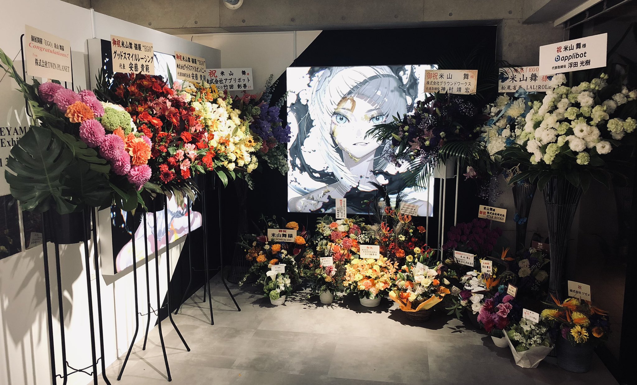 the image shows a room with a black and white animestyle portrait of a woman on the wall. the room is decorated with numerous bouquets and flower arrangements in various sizes and colors.  many of the bouquets have messages written in japanese on them. the lighting is artificial and the overall atmosphere is festive, possibly for a celebration or an exhibition opening.