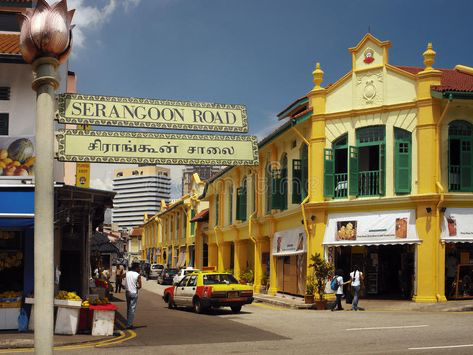 a vibrant street scene in singapore along serangoon road. the image features rows of colorful, traditional shop houses, predominantly yellow, with green window shutters. a street sign in both english and tamil script marks the location as serangoon road. people are walking along the street, and cars are parked and driving. the scene is sunny and bustling, suggesting a lively atmosphere of a local market or commercial area. the architecture reflects a mix of colonial and asian influences, hinting at the rich cultural heritage of the location.  in the background, modern buildings can be seen, showing the juxtaposition of old and new within the city's fabric.