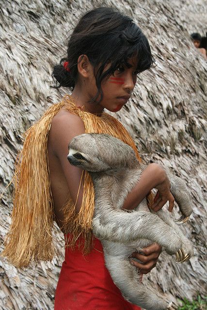 a young indigenous girl from the amazon rainforest holds a sloth in her arms. she is wearing a red dress and traditional clothing made of brown fibers. the girl is smiling and looks happy. the background is a blurred view of the lush rainforest.
