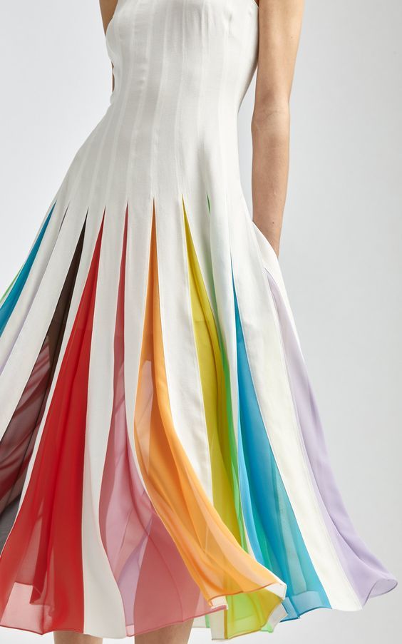 a white, kneelength dress with colorful pleated details is showcased. the dress has a fitted bodice with a high neckline and vertical white panels throughout. the skirt features wide pleats that open to reveal rainbow colored panels including hues of red, orange, yellow, green, blue, indigo, and violet. the movement of the fabric suggests that the skirt is made of a lightweight, flowing material. the model is visible from the chest down, standing against a simple white background.