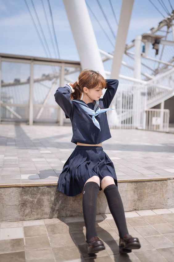 a young woman wearing a japanesestyle sailor school uniform sits on a set of concrete steps. she is tying her hair back with both hands and looking down. she wears black kneehigh socks and black mary jane shoes. behind her is a white metal structure that appears to be a bridge or walkway. the background is blurred, suggesting a bright and sunny day.