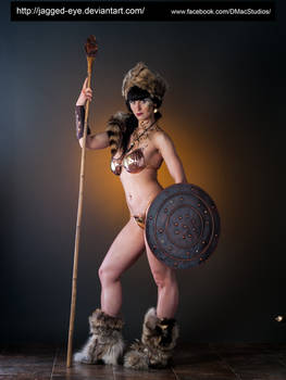 the image shows a female model posing as a fantasy warrior.  she's wearing a  bronzecolored bikinistyle top and a furtrimmed headdress.  her attire also includes  long fur boots. she holds a wooden spear in one hand and a round, dark brown shield in the other. the background is a muted dark brown/gray. the overall style suggests a fantasy or tribal warrior aesthetic. the photo is professionally lit and composed, suggesting a studio photoshoot.  watermarks indicating the photographer's deviantart and facebook pages are present.