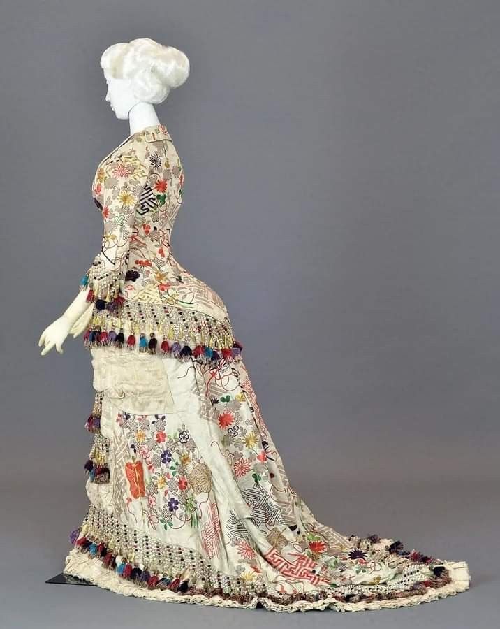 a photograph of a mannequin wearing a vintage dress. the dress is white and features floral embroidery. it also has tassels around the hem. the mannequin has a white wig. the background is gray.