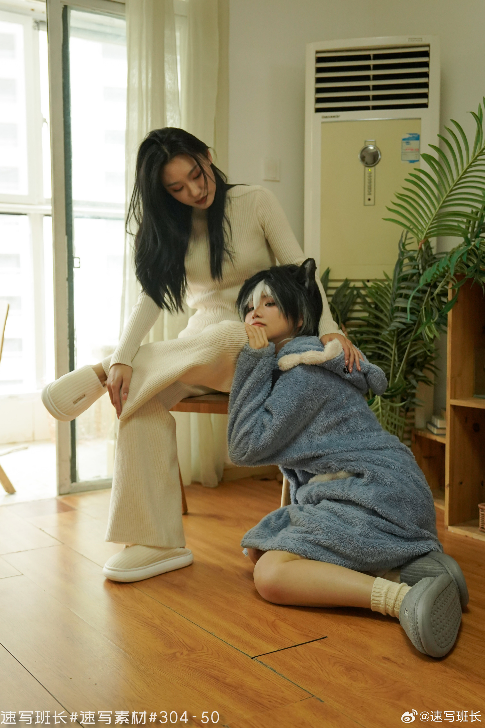 two women are posing for a photo shoot. the woman on the left is wearing a white sweater and pants, while the woman on the right is wearing a blue cat costume. the woman on the left is sitting on a chair, while the woman on the right is kneeling on the floor. they are both looking at the camera. the photo was taken in a room with wooden flooring and white walls. there is an air conditioner in the background.