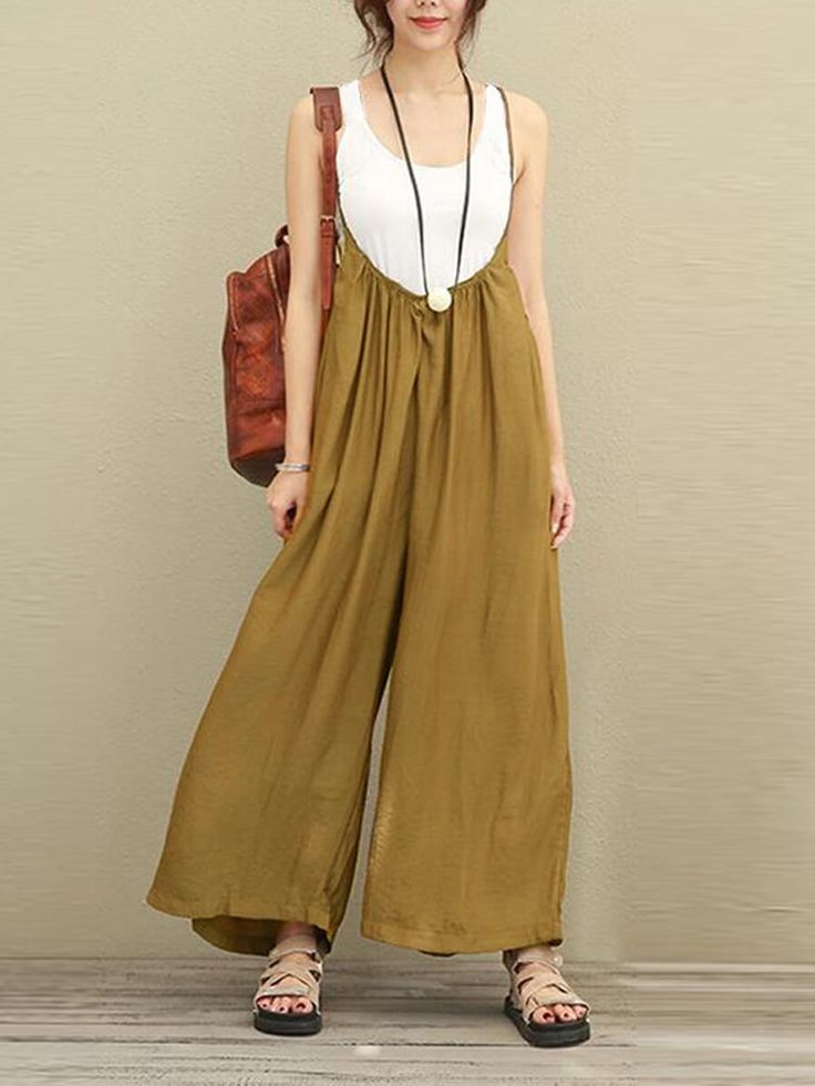 a fulllength shot shows a woman wearing a light olive green jumpsuit with a white tank top underneath. she has a brown backpack slung over her left shoulder and is wearing sandals. a long black necklace with a round pendant hangs down to the center of her chest.
