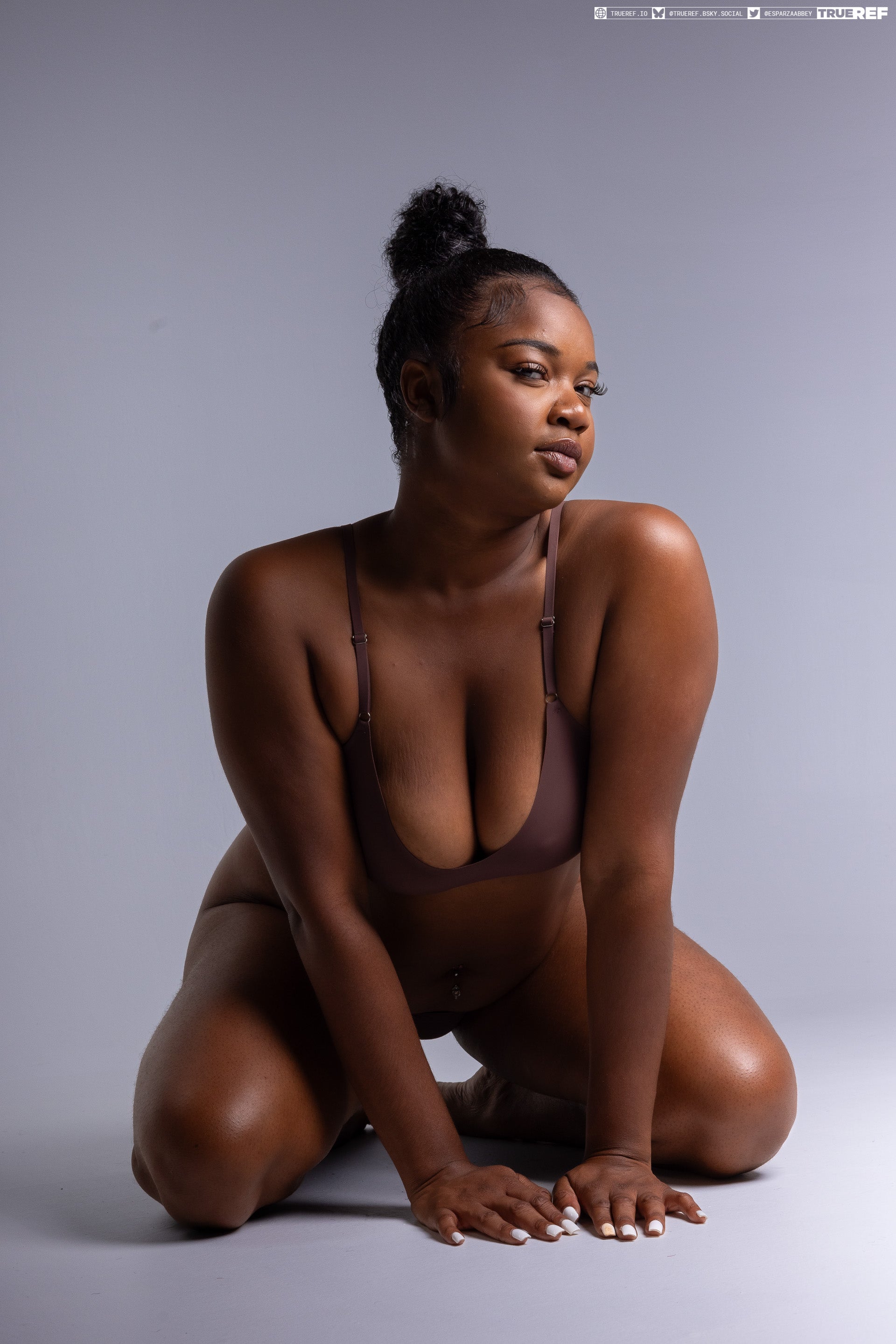 a portrait of a darkskinned woman kneeling in a studio wearing a brown bra with her hair in a bun.