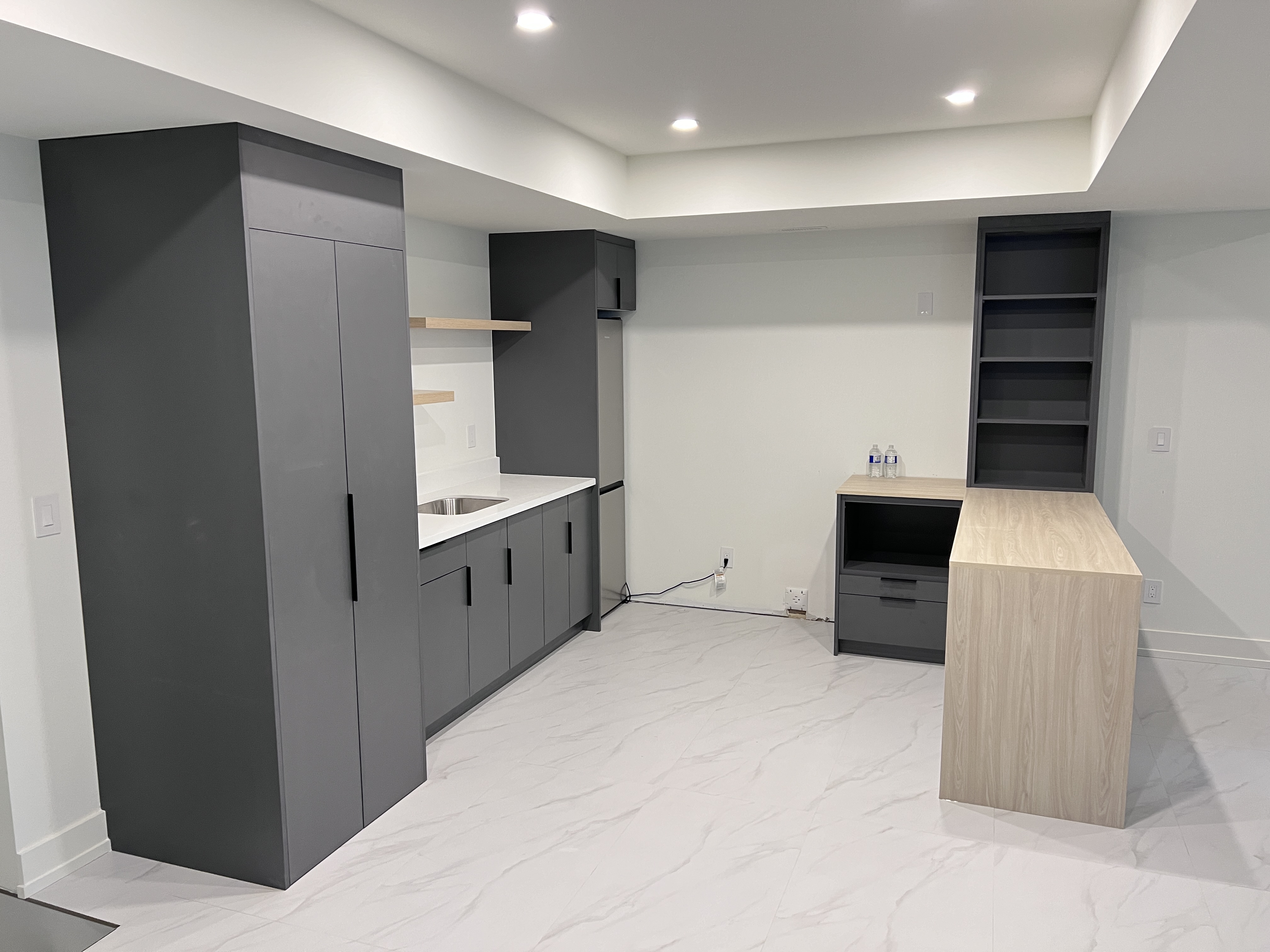 a modern kitchen with gray cabinets, white countertops, and a wood countertop. the kitchen has a sink, refrigerator, and shelves. the floor is white tile. there is a large window on the left side of the room. the walls are painted white. the kitchen is clean and modern.