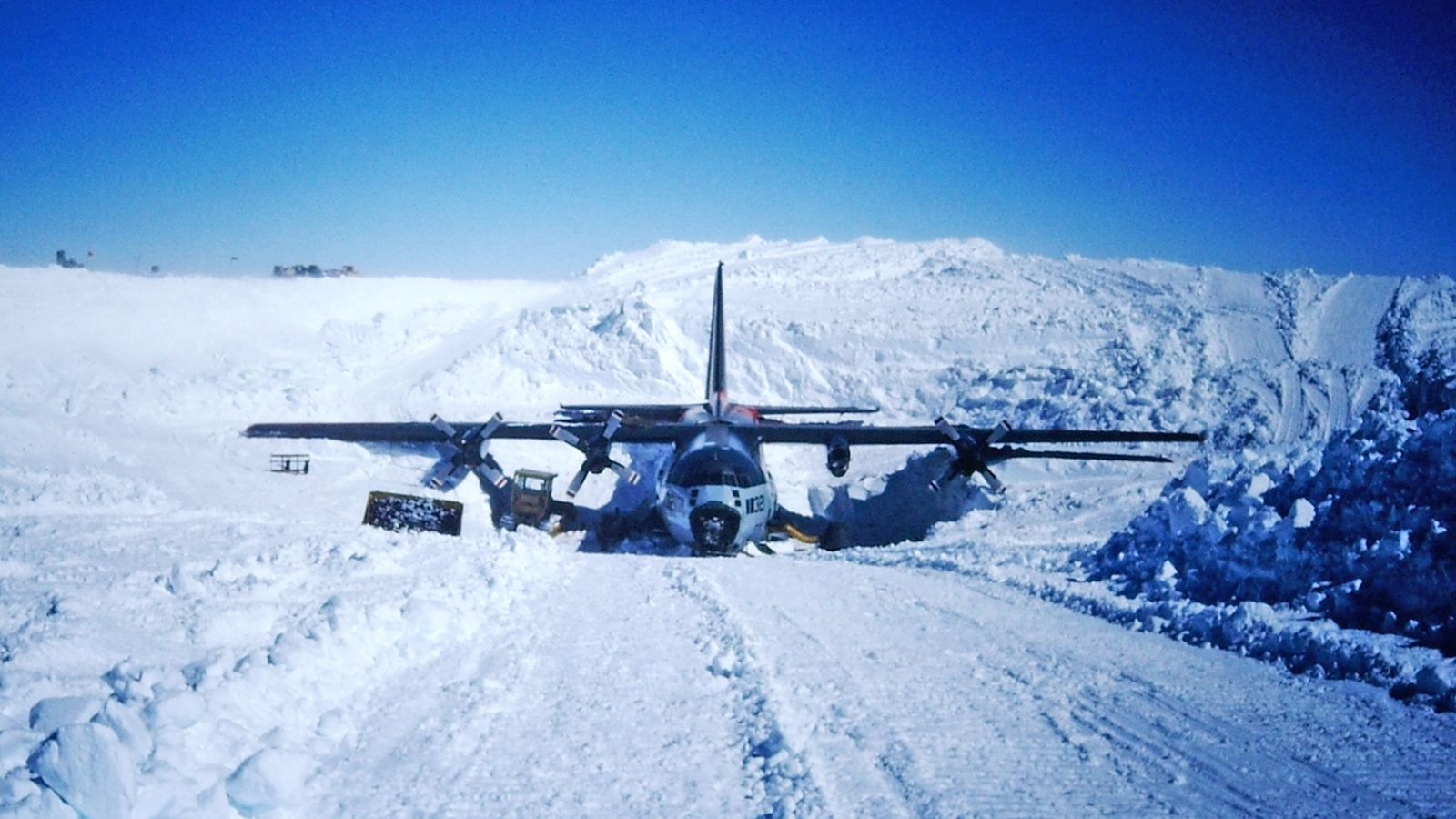 a military cargo plane sits on a snowy landscape, its landing gear firmly planted on the frozen ground. the plane is surrounded by deep snowdrifts and white mountains, creating a stark and beautiful backdrop. the clear blue sky above adds to the sense of isolation and grandeur.