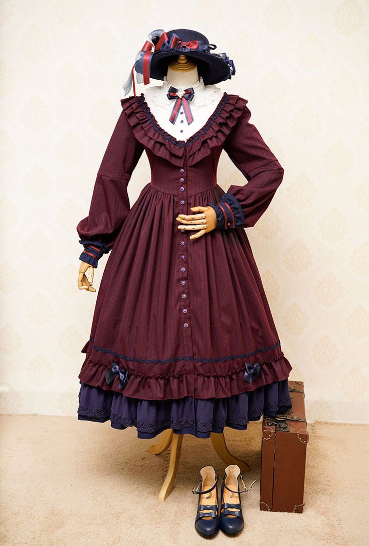 a mannequin dressed in a maroon lolita fashion dress, complete with a matching hat and blue shoes. the dress features intricate lace detailing, a ruffled collar, and a tiered skirt. a vintage suitcase is placed next to the mannequin, adding to the vintage aesthetic. the image is taken indoors with studio lighting, providing a clear and welllit view of the outfit.