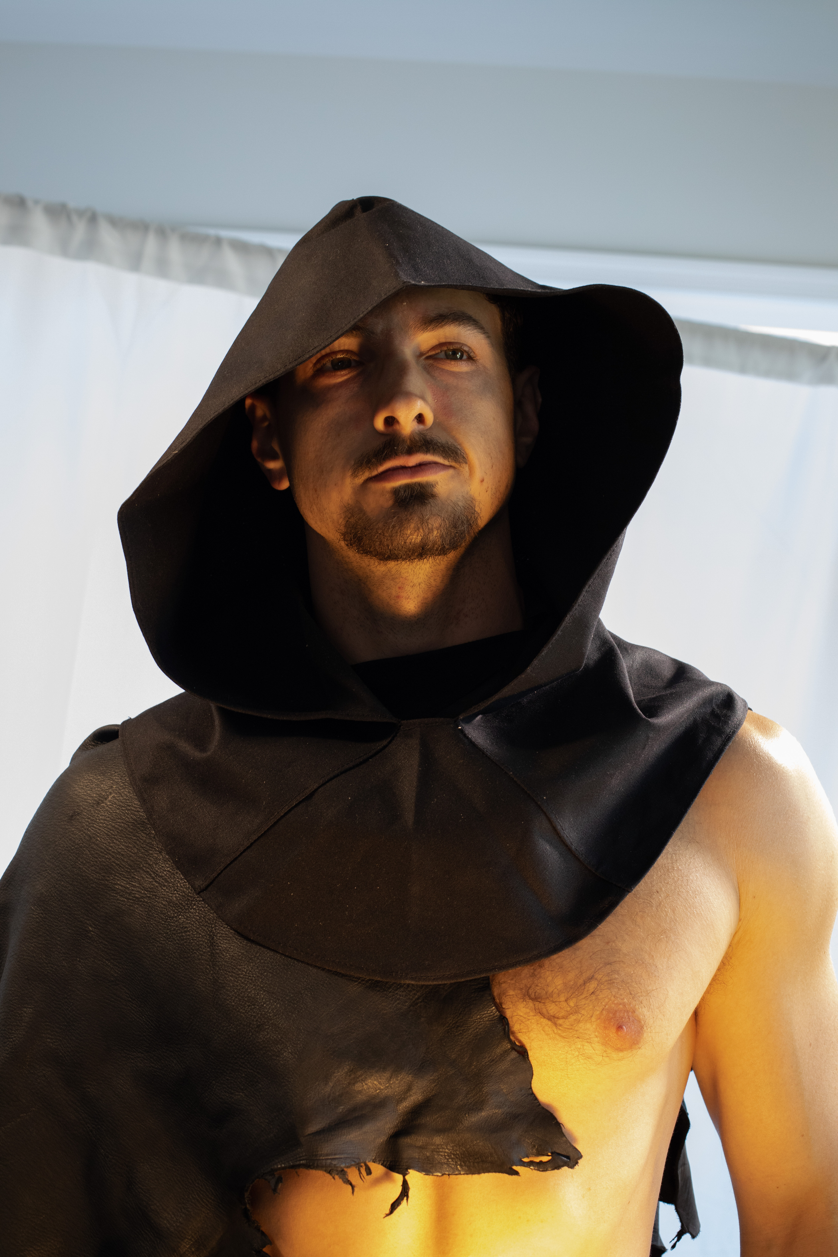 a man with an intense gaze wears a dark hooded cloak, partially revealing his bare, muscular chest. the cloak is torn, suggesting experience and perhaps battle. the lighting is dramatic, highlighting his features and creating a sense of mystery and power.