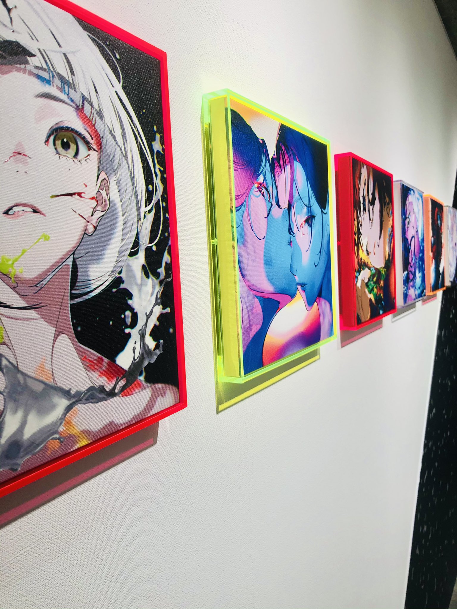 the image shows a series of squareframed anime artworks mounted on a white wall. the artworks are brightly colored and feature female characters. the frames are illuminated by artificial lighting, casting a slight shadow on the wall. the perspective is from the side, offering a glimpse into the art exhibition.