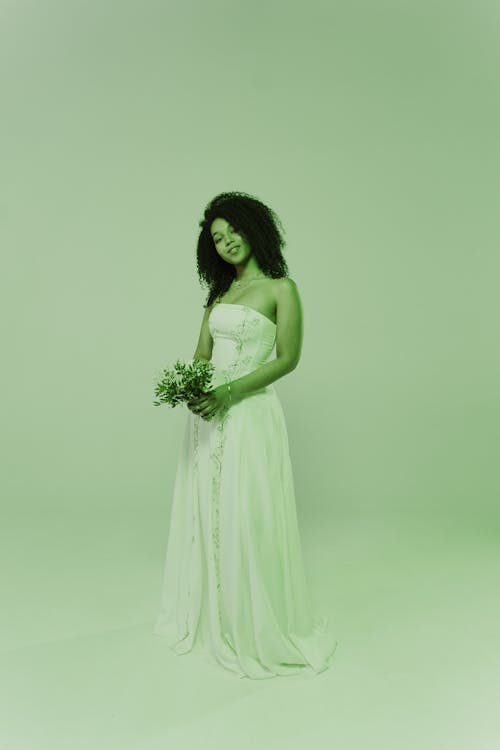a fulllength shot of a beautiful black bride in a white wedding dress. she is standing against a pale green background, holding a small bouquet of flowers.  she has curly hair and appears happy and serene. the overall tone of the image is soft and romantic.