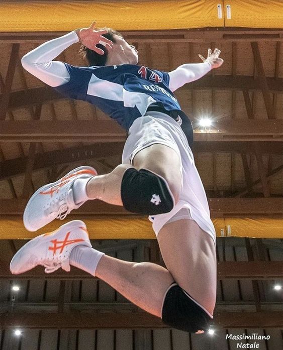 a male volleyball player is captured midair as he executes a powerful spike. he is wearing a blue and white jersey, white shorts, and knee pads. the perspective is from below, emphasizing the height and power of his jump. the setting is a gymnasium with wooden flooring and bright overhead lighting.