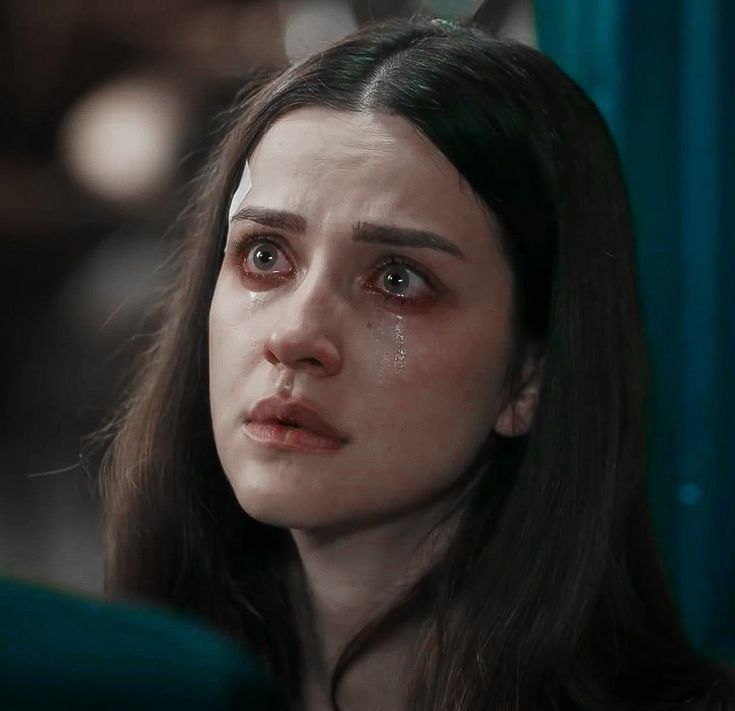 a woman with dark hair and a bandage on her forehead is crying. she is looking up and to the left of the frame. her eyes are red and swollen, and tears are streaming down her face. the background is dark and out of focus.