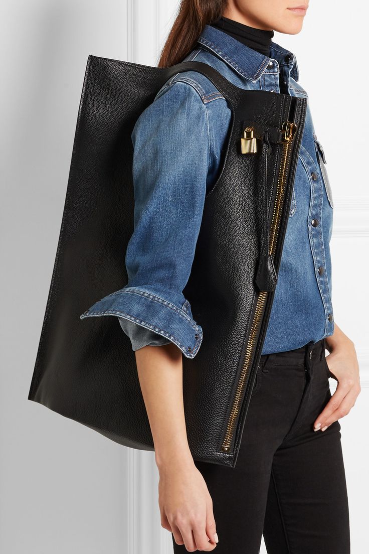 a woman is shown from the waist up, wearing a denim shirt over a black turtleneck, and black jeans. she is carrying a large, rectangular black leather tote bag over her shoulder. the bag has a goldcolored zipper along its side, and a small gold padlock attached near the top. the woman's arm is at her side, and she is looking slightly away from the camera. the background is a plain white wall.