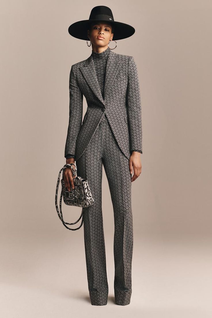 a fashion model, a woman, is posing in a trendy suit, a black and white patterned jacket and trousers, a black hat and a white handbag. she is standing with her hands on her hips.  the background is a simple beige, allowing the clothes to be the focus.