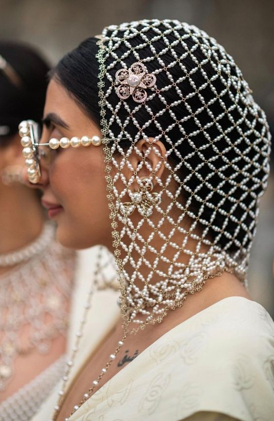 a closeup shot of a woman with a pearl veil and sunglasses in a white dress. the veil is made of small white pearls and is arranged in a netlike pattern. the woman's hair is dark and styled with the veil.  she is wearing a pair of white pearl sunglasses. the veil and sunglasses create a very elegant and stylish look. the woman's face is in focus, and her expression is calm and relaxed.