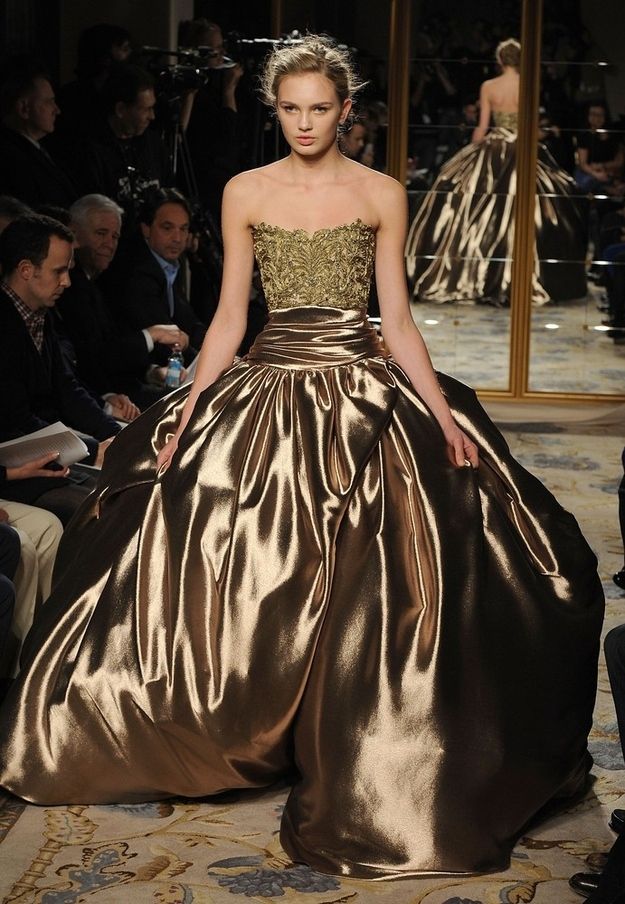 a beautiful woman wearing a long gold ball gown poses on the runway during a fashion show. the gown is made of silk satin and features a fitted bodice and a full skirt. the model is wearing her hair up and has a natural makeup look. she looks elegant and glamorous as she walks the runway.