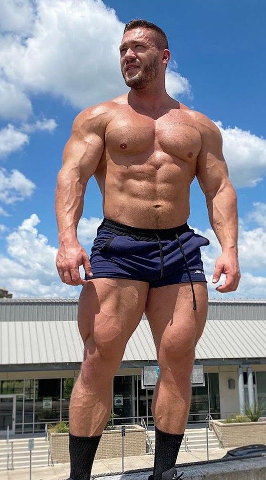 the image shows a muscular, tanned man with a welldefined physique. he is shirtless, wearing navy athletic shorts, and black ankle socks. his body displays significant muscle mass and definition, including prominent sixpack abs and large, strong legs. he is standing outdoors on a sunny day with a partly cloudy sky in the background. behind him is a modernlooking building. the man appears confident and is possibly posing for a photo. the overall impression is one of fitness, strength, and health.