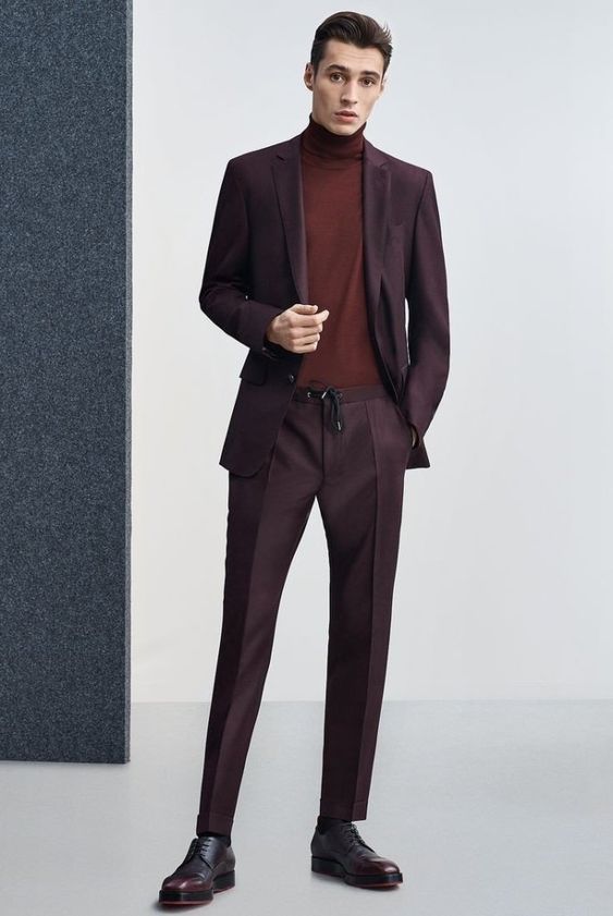 a full shot of a man in a maroon suit and turtleneck sweater. he is standing and posing indoors with his right hand tucked inside his jacket. he is wearing dark shoes and the pants appear to have a drawstring waist. a dark gray textured wall is on the left side of the image.