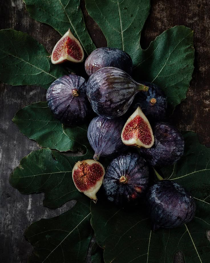 the image shows a still life arrangement of figs and fig leaves on a dark wooden surface. several whole figs with purple skins are clustered together among the green fig leaves. three figs have been cut in half, revealing their red interiors. the overall tone is dark and moody.
