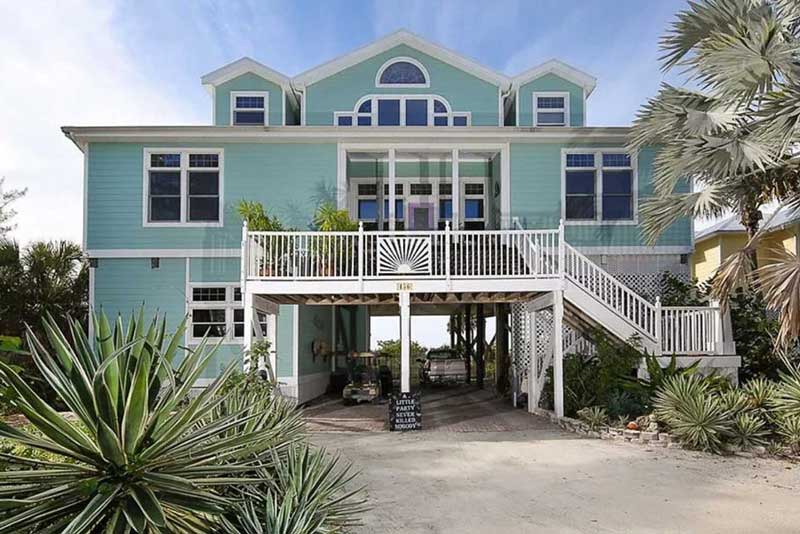 a tealcolored stilt house with white trim stands out against a clear blue sky.  the home features a large porch on the second story, accessible by two sets of stairs.  lush tropical plants adorn the ground level, flanking the driveway.  a glimpse of a yellow house is visible to the right.  the overall feel is bright, cheerful, and welcoming.