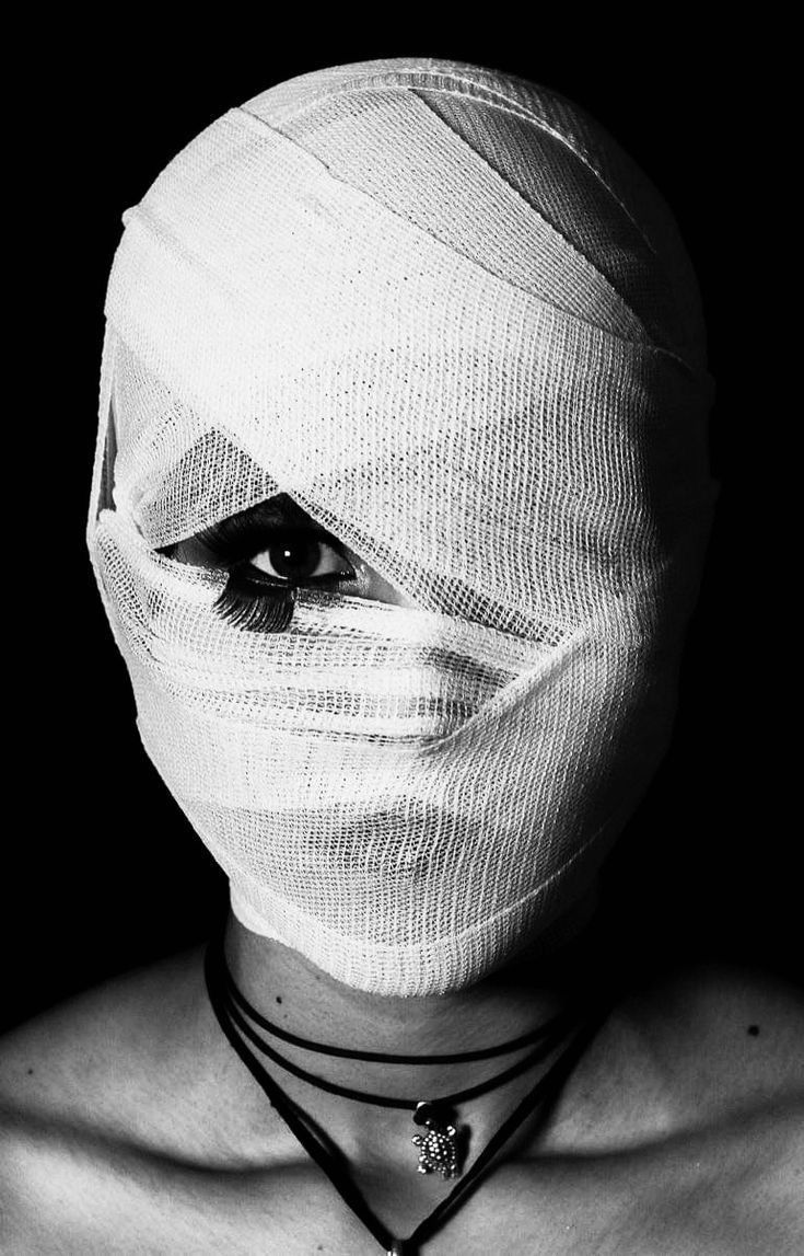 a black and white closeup portrait of a woman's face wrapped in a white bandage. only one eye with dramatic makeup is visible. she is wearing a simple necklace with a turtle pendant. the image is dark and dramatic, creating a mysterious and slightly unsettling mood. the contrast between the stark white bandage and the dark background emphasizes the subject's face and the single visible eye. the overall style is artistic and fashionforward.