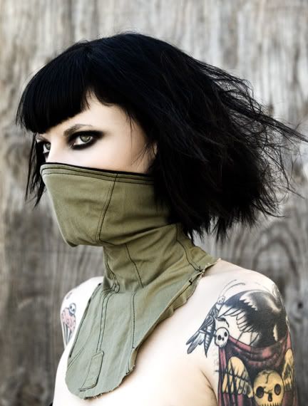 a woman with black hair and dark eyes stares intensely at the camera. she wears a green, fabric collar and a black shirt. the woman has multiple tattoos on her shoulders and arm, including a bird and skull design on her right shoulder. the woman is in focus, while the background of wood is out of focus. her serious gaze and tattoos give her a dark and mysterious feel.
