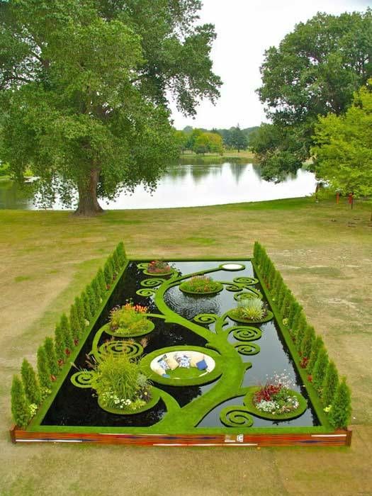 this image features a meticulously designed formal garden with a black pond as its centerpiece. the pond is decorated with green, grasscovered spiral patterns that form floating islands with various plants and flowers. the garden is bordered by neatly trimmed hedges of topiary and lush green grass. in the background, there is a scenic view of a lake surrounded by trees, creating a beautiful natural landscape.