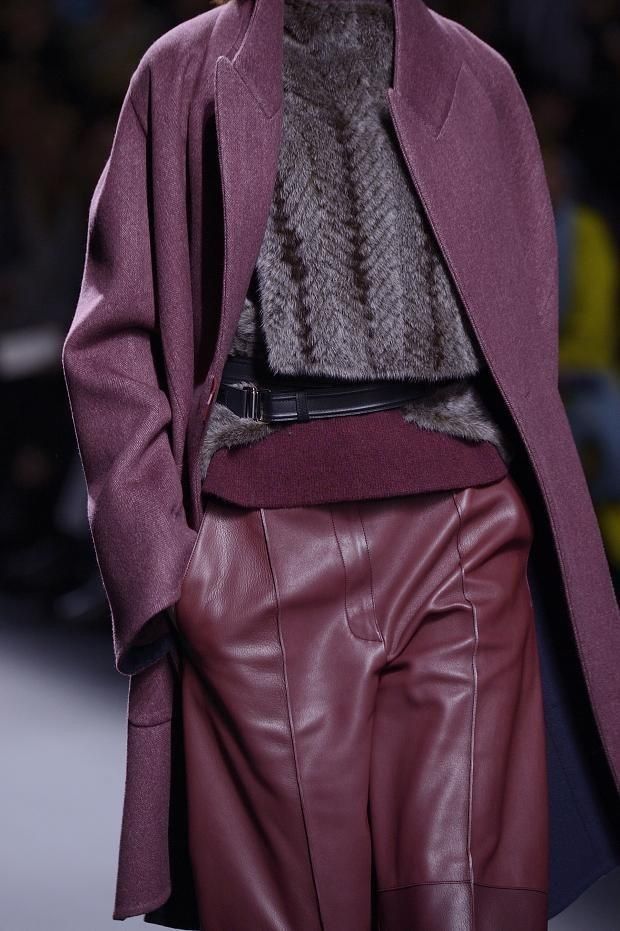closeup shot of a female model showcasing a stylish outfit on a runway. the outfit consists of a long, burgundy wool coat, a grey herringbone patterned sweater, and burgundy leather pants. a black belt cinches the waist. the focus is on the texture and color coordination of the clothing items. the background is blurred, keeping the attention on the model's ensemble.  the overall impression is one of sophisticated and chic autumn/winter fashion.