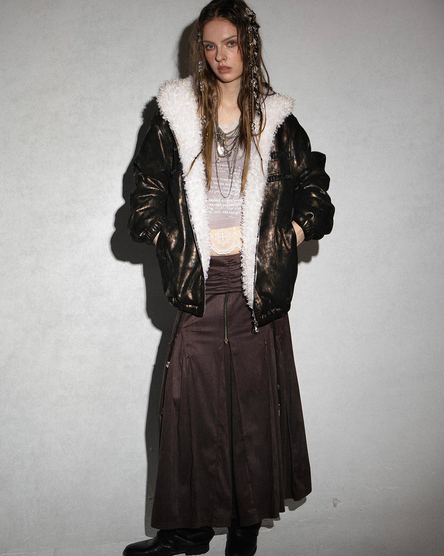 a fullbody studio shot depicts a young woman modeling a stylish autumn/winter outfit.  she is wearing a dark bronze metallic jacket with a white sherpa lining and hood. underneath, she wears a long, dark brown aline skirt with pleats and a front zipper detail. her accessories include multiple necklaces and her hair is styled in long braids. the overall style is a mix of grunge, bohemian and edgy elements. the backdrop is a simple light grey wall.  she is wearing dark boots.