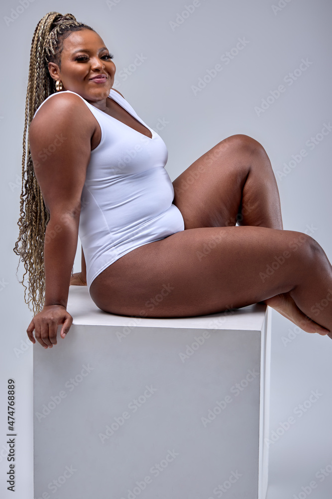 a beautiful black woman sits on a white platform. she is wearing a white bodysuit and has her legs crossed. she is smiling and looks confident and happy. she is a plus size model and is embracing her curves.