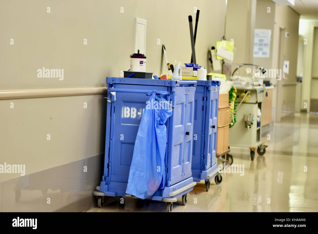 two blue cleaning carts with supplies are parked in a hospital hallway. the carts are filled with cleaning supplies, including mops, buckets, and cleaning solution. the hallway is empty, and the floor is clean and shiny. the walls are painted white.  the carts have a blue plastic covering to prevent crosscontamination. this is a common sight in hospitals and other medical facilities, as cleanliness is of paramount importance.  the lighting in the hallway makes the floor appear glossy.