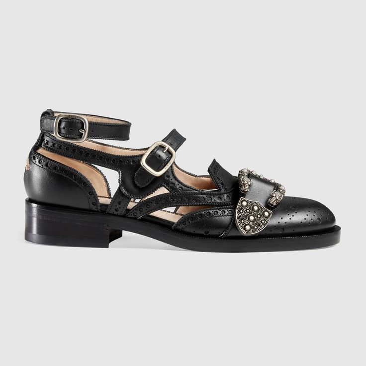 the image shows a single black leather women's shoe from gucci.  the shoe is a brogue style with cutouts and perforations throughout the leather upper. it features an ankle strap with a buckle closure, and additional buckles adorned with metal studs on the vamp.  the shoe has a slightly raised platform sole and a rounded toe. the overall style is a blend of classic brogue elements with modern cutouts and embellishments.  the background is plain grey, providing a neutral backdrop that highlights the shoe.