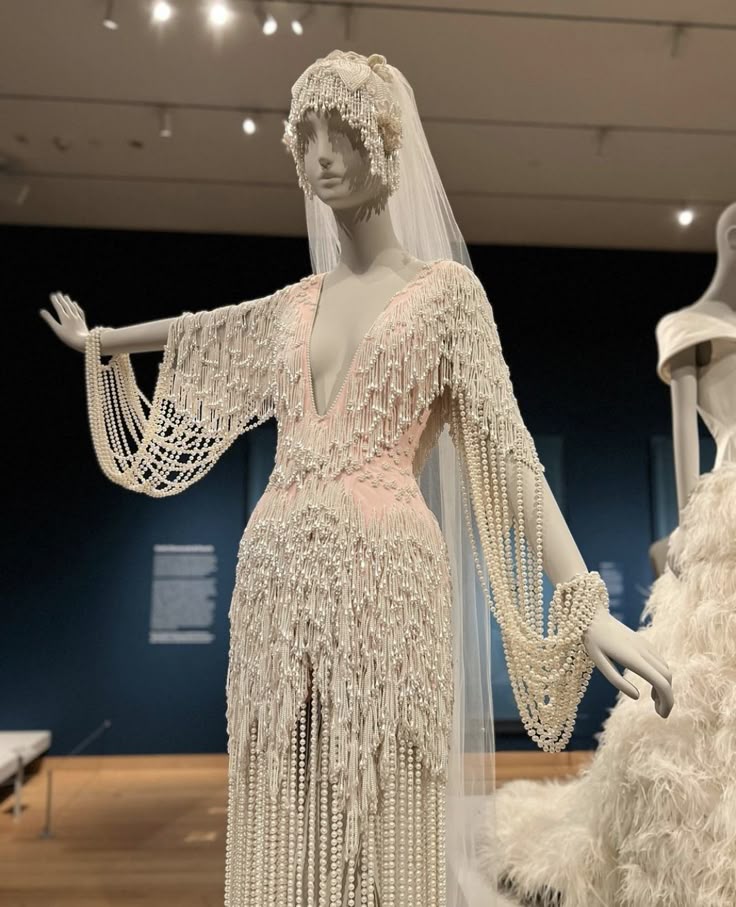 a mannequin wearing a light pink and white beaded wedding dress with a deep vneck, long beaded sleeves, and fringe is on display in a museum. the mannequin is also wearing a matching beaded headpiece and veil. another wedding dress is partially visible on the right side. the background includes a blue wall and a wooden floor.