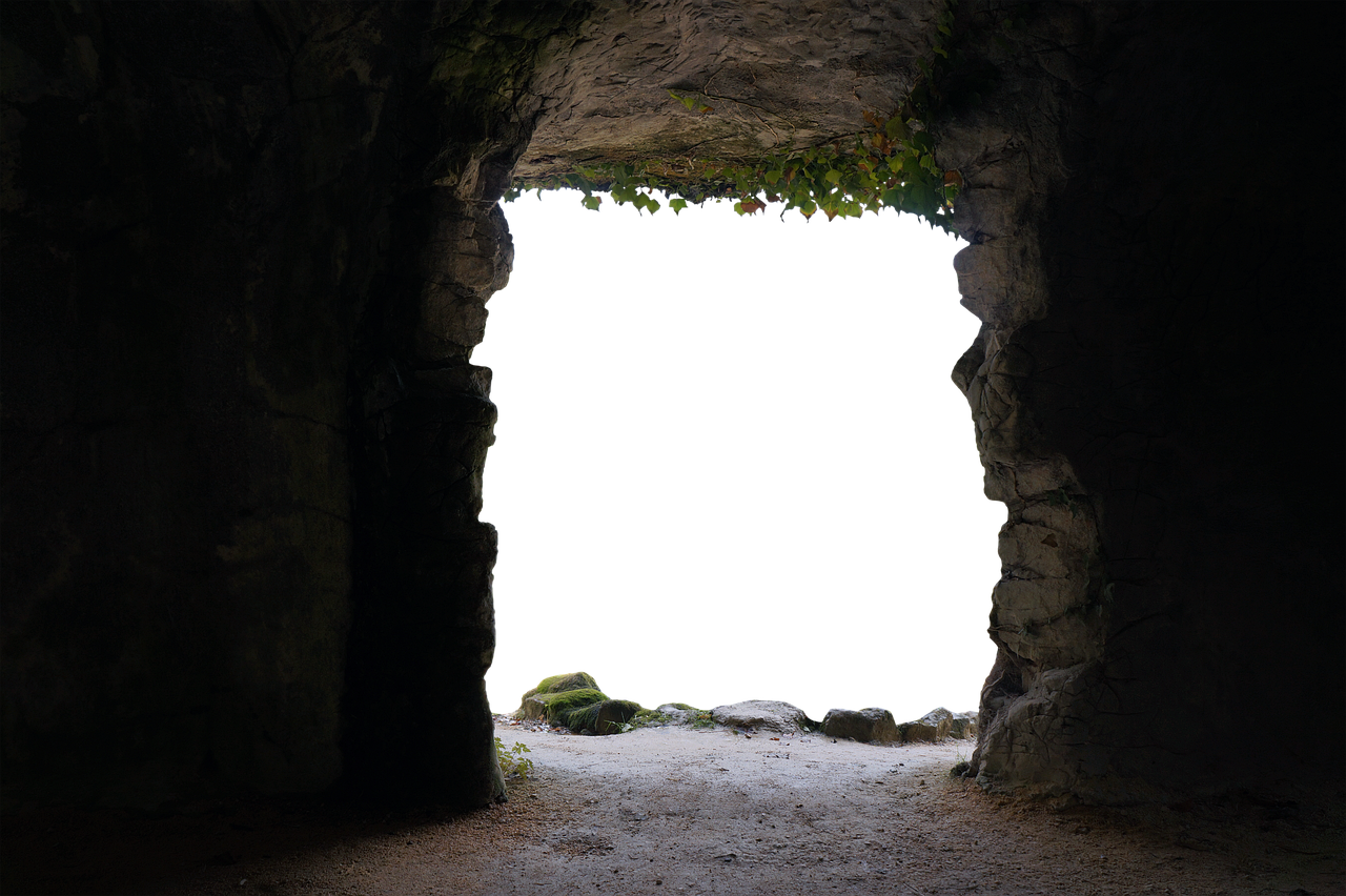 the image shows a dark cave entrance framed by rough rock formations. the opening is a deep black, contrasting with the lighter, textured stone of the cave walls. moss grows on some of the rocks, adding a touch of greenery. the floor is a mixture of dirt and small stones, leading into the darkness beyond.