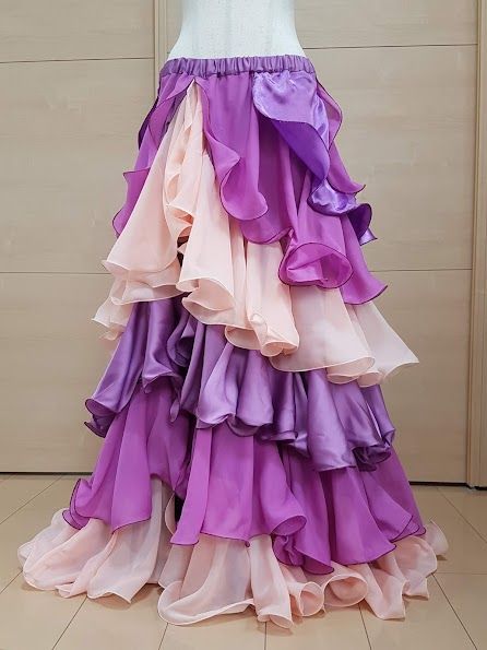 a tiered, ruffled skirt in shades of purple and pink is displayed on a mannequin. the skirt has an elastic waistband and is made of a lightweight fabric, possibly chiffon. the tiers are arranged in alternating colors, with ruffles adding volume and movement to the skirt. the background is a neutral wall and floor.