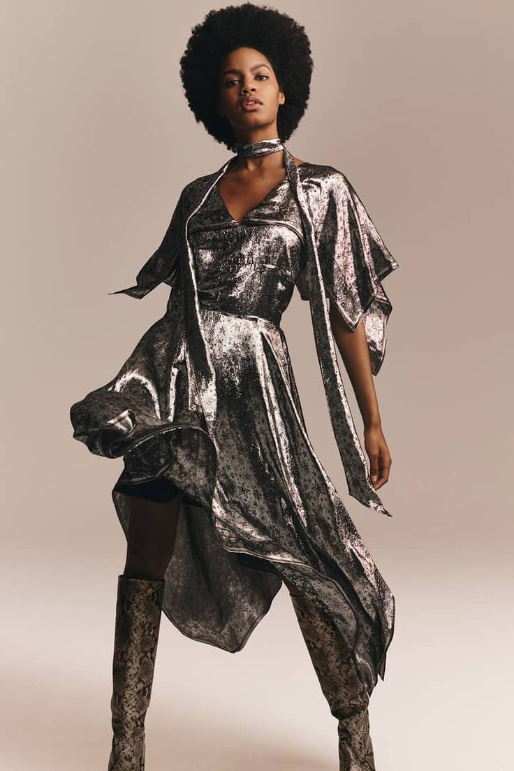 a beautiful young black woman with an afro is posing for a fashion shoot. she is wearing a stunning silver metallic dress with a  snake print. the dress is asymmetrical and has a flowing silhouette. she is also wearing matching snake print boots that add to the glamour of the look. the model's confidence and poise shine through in this powerful image.