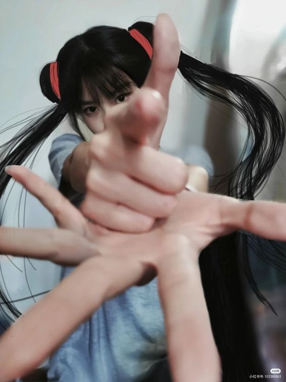 a young woman with long black pigtails in a grey tshirt is taking a selfie with her hand reaching out towards the camera.  she is indoors and the lighting is soft.