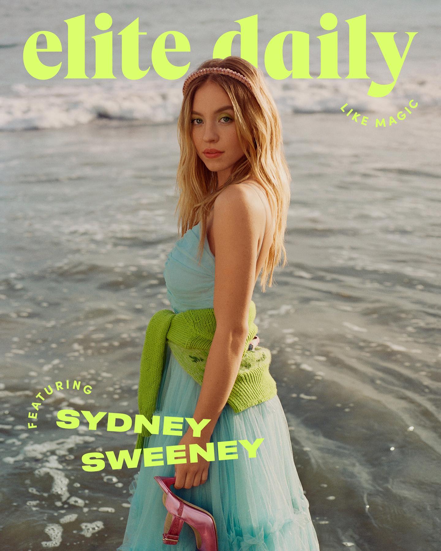 the image is a magazine cover for elite daily featuring actress sydney sweeney.  sweeney is posed on a beach, the water lapping at her feet. she's wearing a light blue, almost pastel, tulle or chiffon dress with a lime green cardigan draped over her shoulders. she has a delicate pink headband in her long blonde hair.  she holds a pair of pink highheeled shoes. the background is blurred but shows the ocean and sky suggesting a sunny day. the elite daily logo is prominently displayed at the top, along with the phrase "like magic" in smaller text.  a circular graphic with "featuring sydney sweeney" is placed in the lower left. the overall mood is bright, airy, and summery.