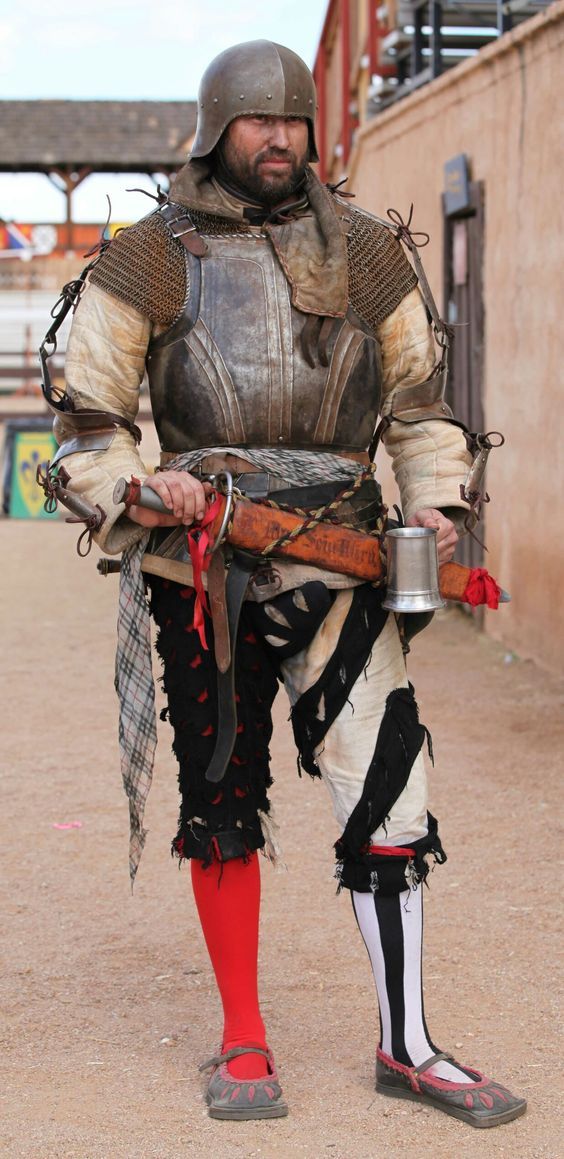 a man wearing a medieval costume with armor, a helmet, a sword, and a dagger stands outdoors.  he is holding a mug and looking at the camera.