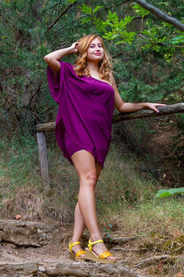 a young, attractive redhead woman stands on a path in a forest, wearing a purple oneshoulder dress and yellow wedge sandals. she is smiling and looking towards the camera. she has her hand gently on her hair and the other hand resting on a wooden fence. the background is lush and green, showcasing a natural outdoor setting.  the overall mood is one of carefree summer beauty.